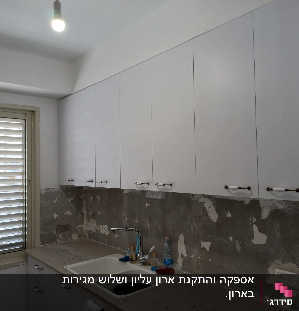 מטבח עם ארונות לבנים, כיור וברז, קיר מתקלף